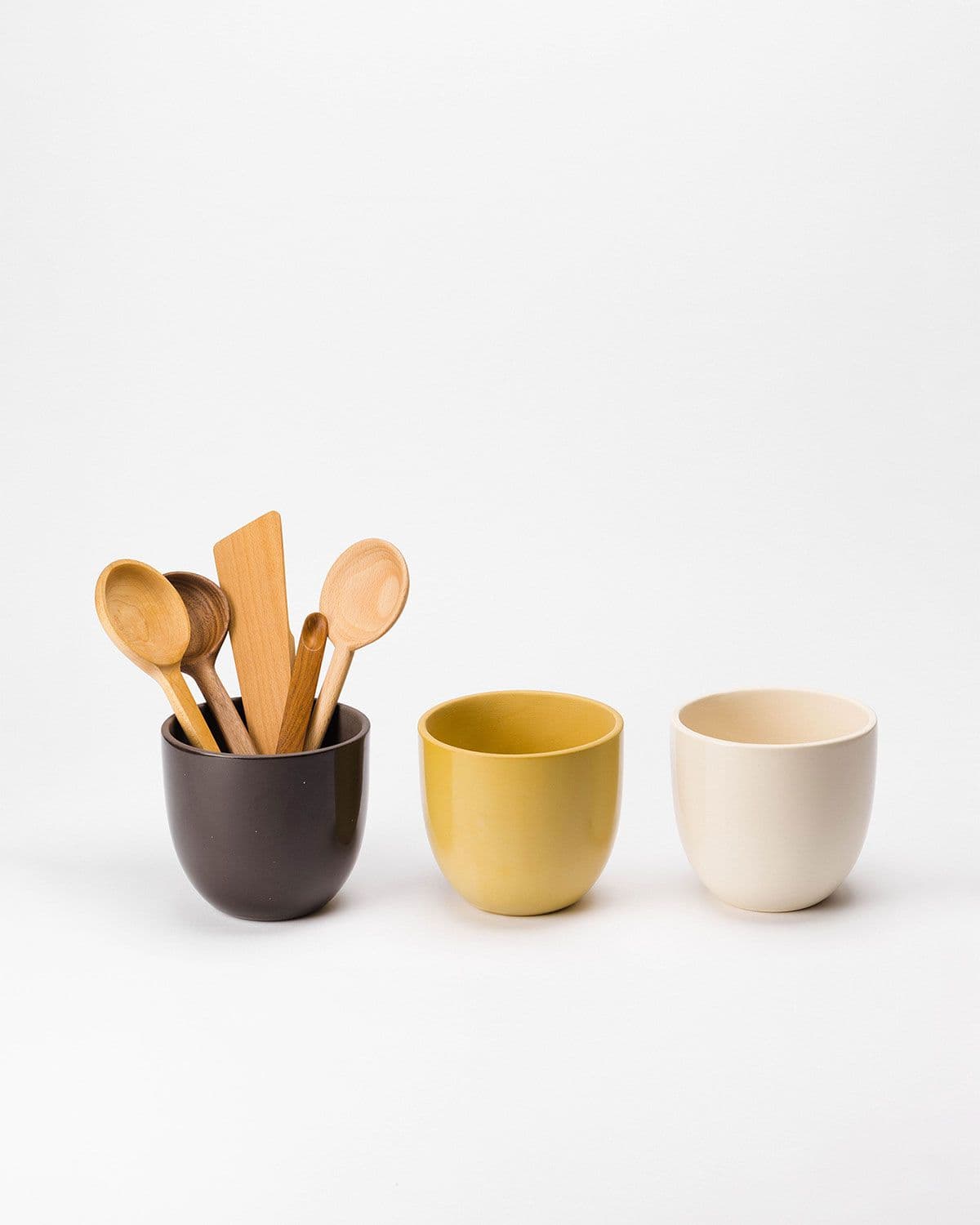 Three ceramic utensil crocks sit in a row upon a white backdrop, left to right: Woods (brown), Pollen (yellow), and Natural (cream). The brown utensil crock on the left holds various wooden utensils.
