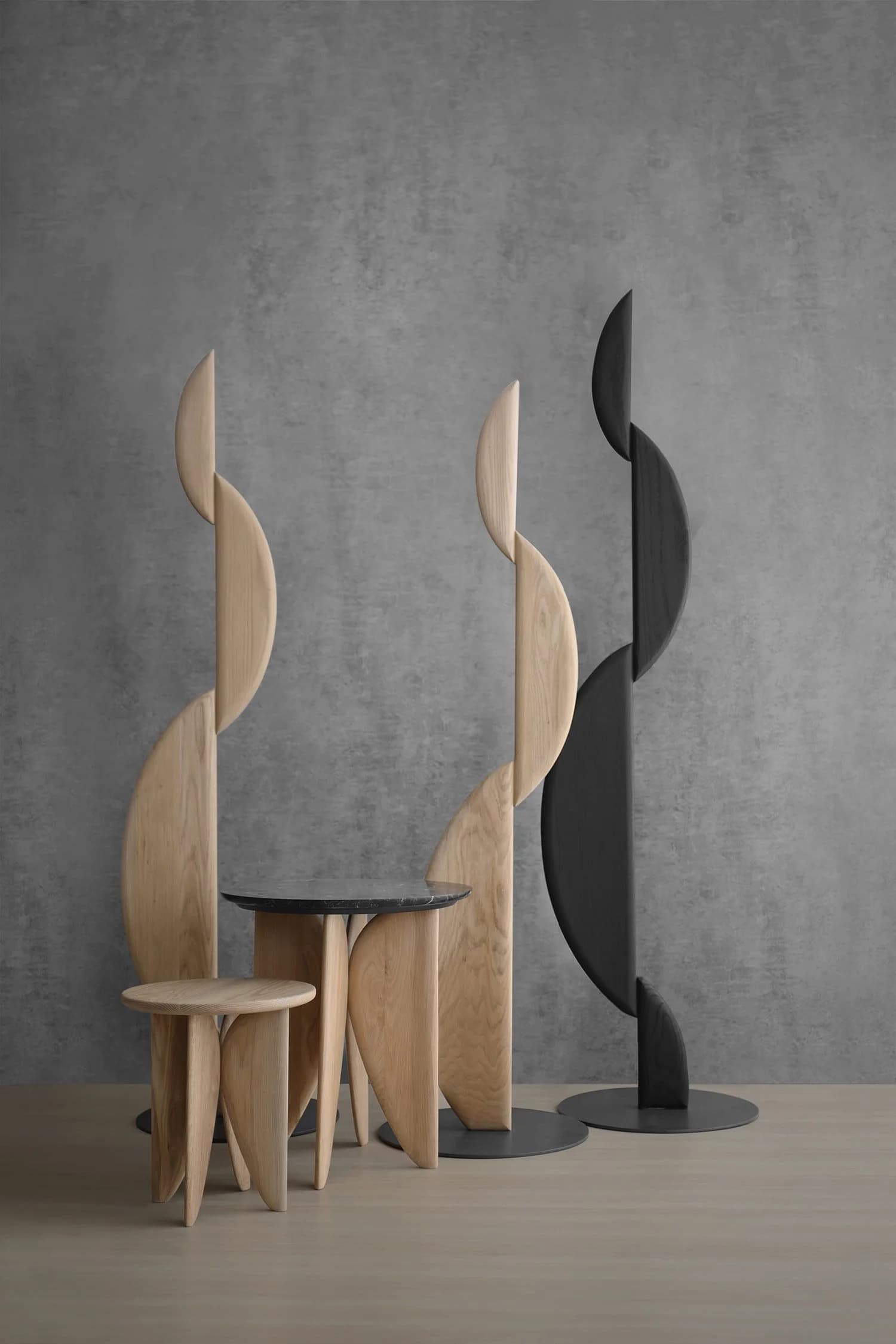 Set of modern sculptural chairs and a small table with a round black top, featuring curved elements resembling abstract leaves, in natural wood and black finishes against a textured gray wall.