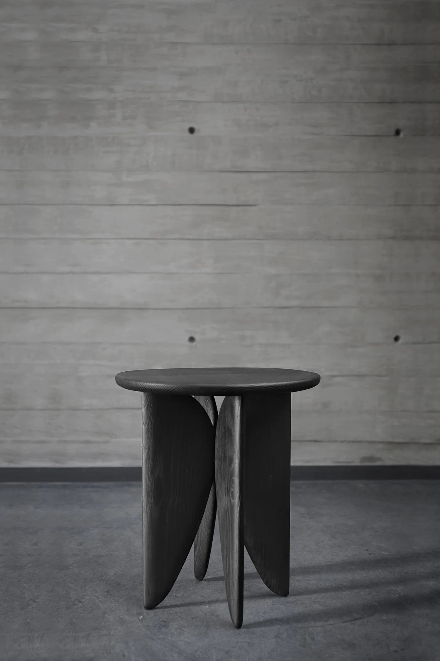 A small, modern black wooden stool with curved legs, set against a gray wooden wall and gray floor.