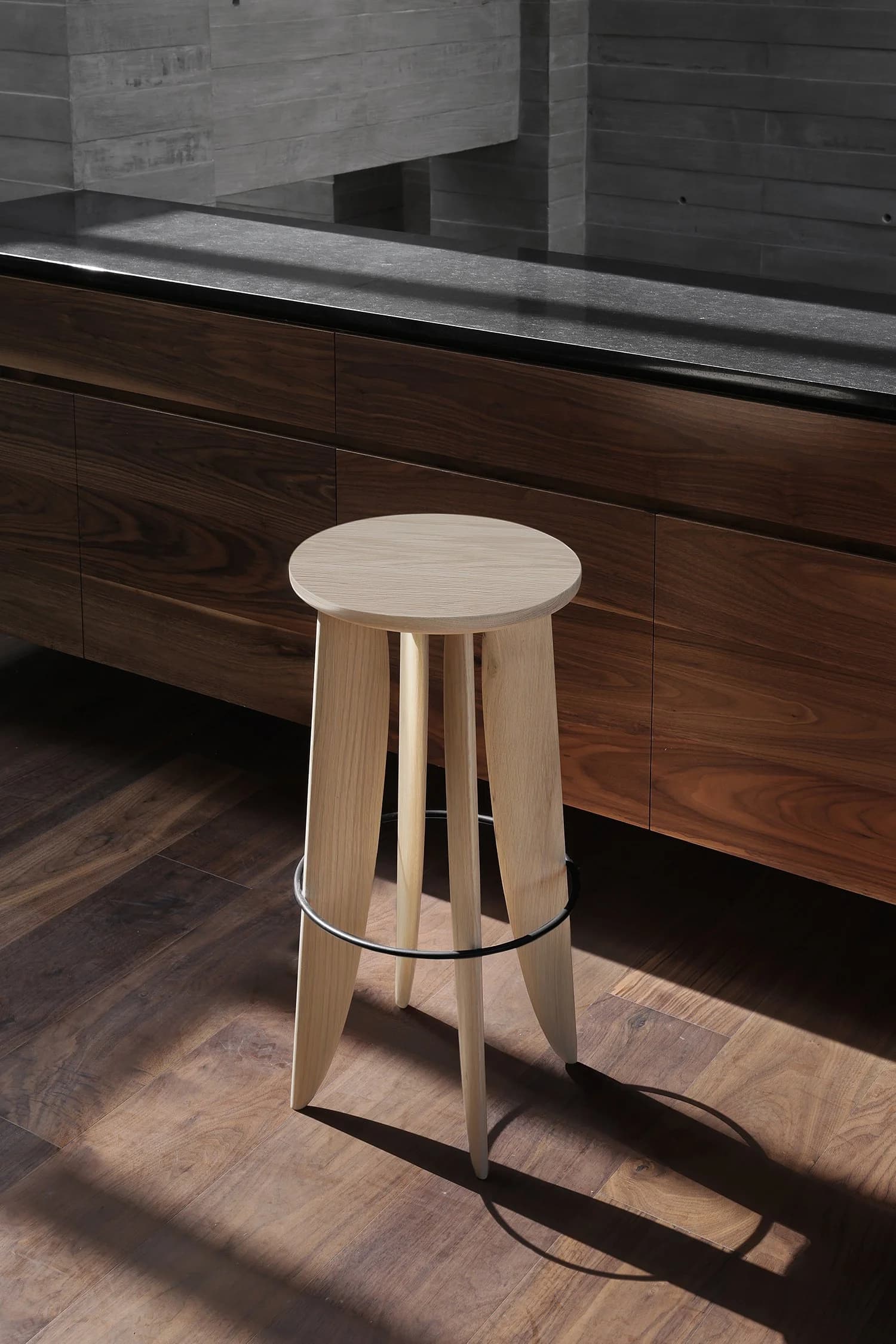 A wooden barstool with four legs and a circular top, placed on a hardwood floor in front of a dark wood cabinet and a black countertop.
