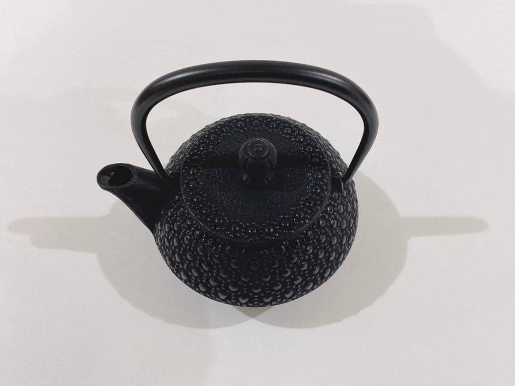 An overhead three-quarter view of a black cast-iron teapot with a textured repeating surface pattern, matching lid and arched handle.