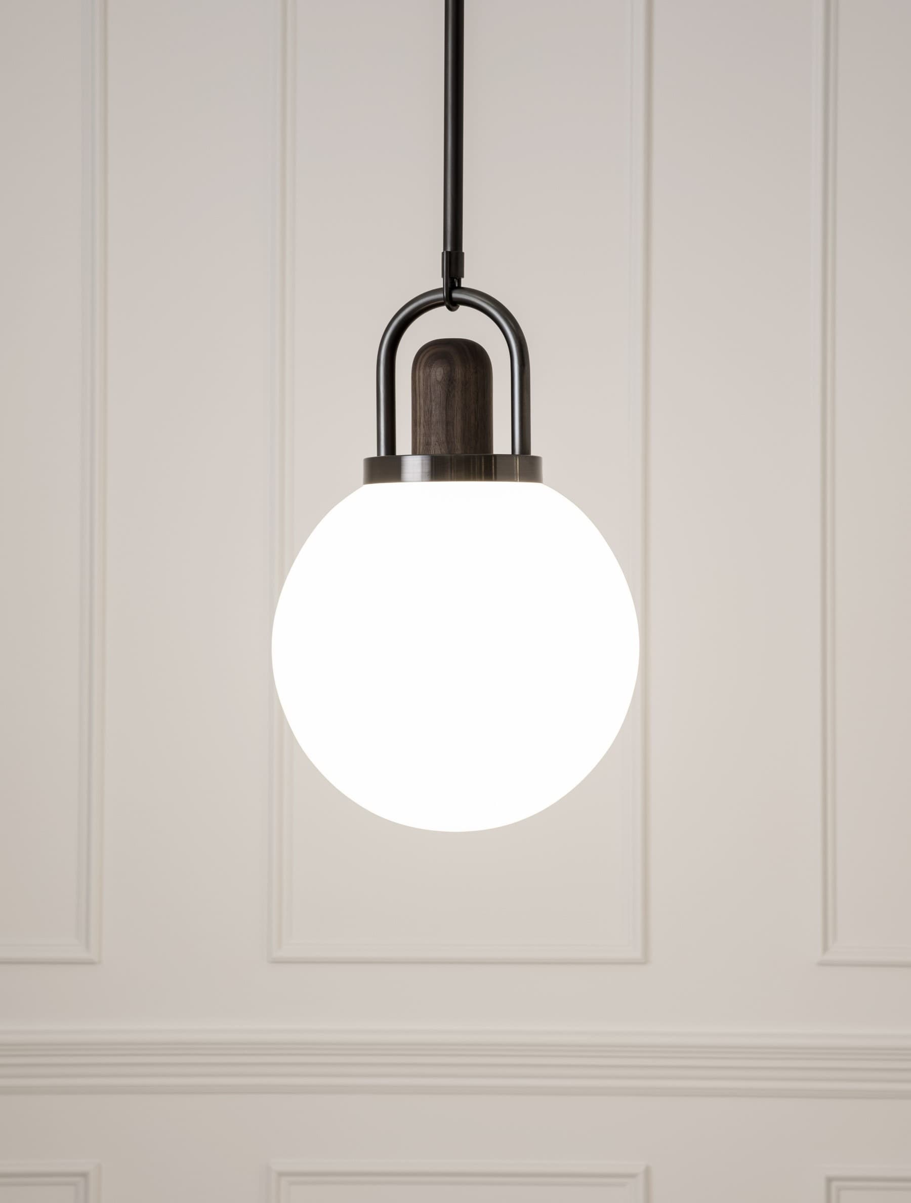An illuminated white glass globe pendant with a dark walnut top inside a blackened-brass arch, against a paneled wall.