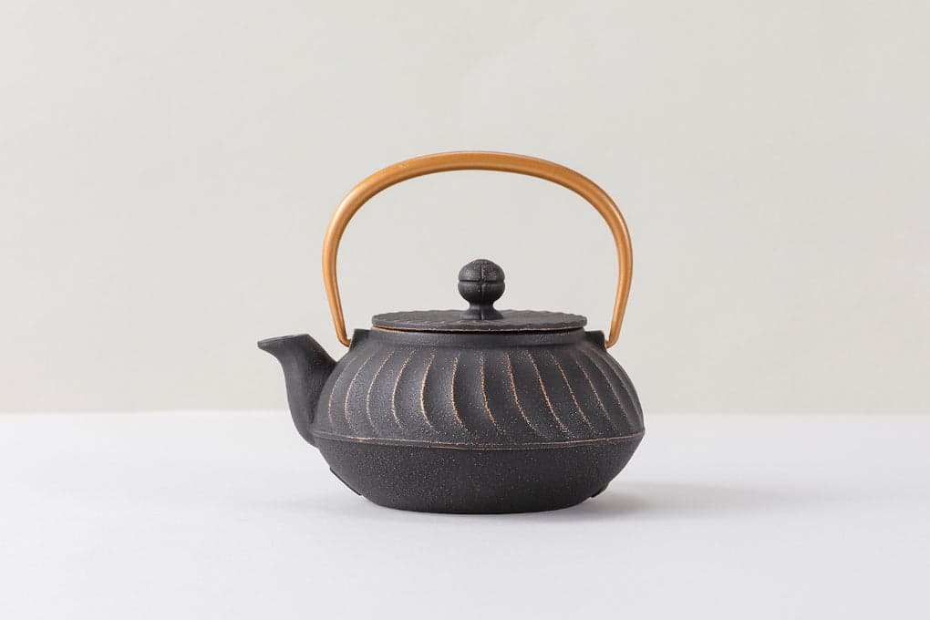 A black cast-iron teapot with gold-highlighted curved wave ridges and a gold-colored arched handle, shown on a pale tabletop.