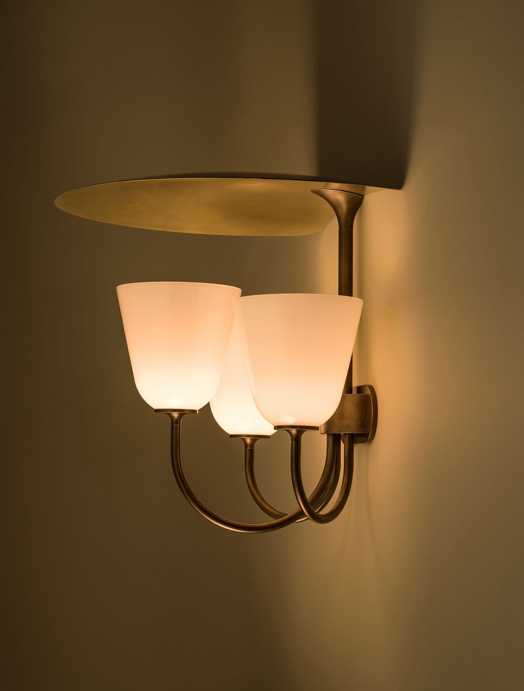 A bronze-finished wall sconce with three glowing opal-glass cups below a broad oval metal reflector.