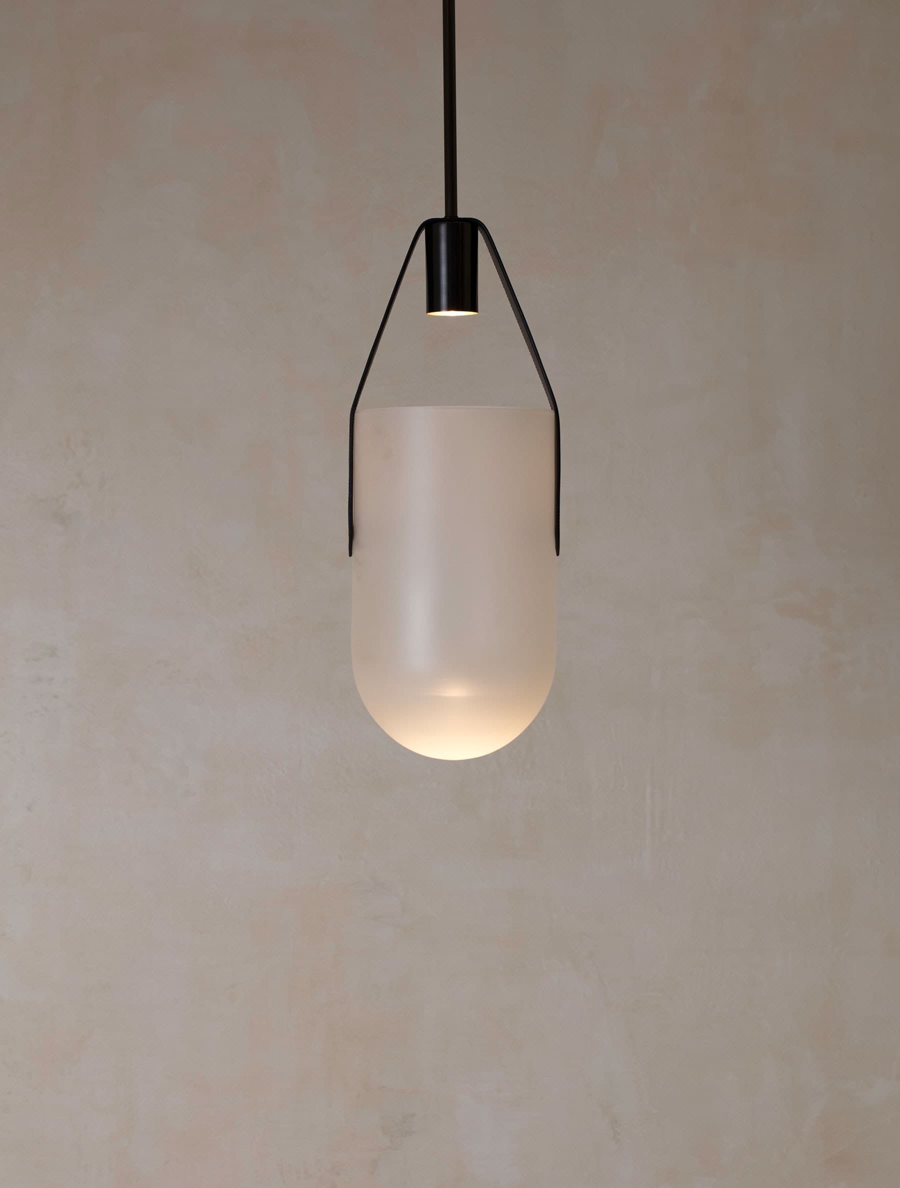 A frosted-glass pendant vessel suspended in black leather straps beneath a small blackened-brass spotlight.