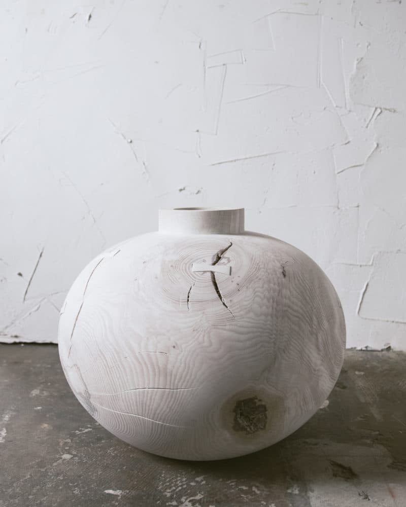A pale rounded wood vessel with a short neck, natural grain and a repaired split stands on a concrete floor.