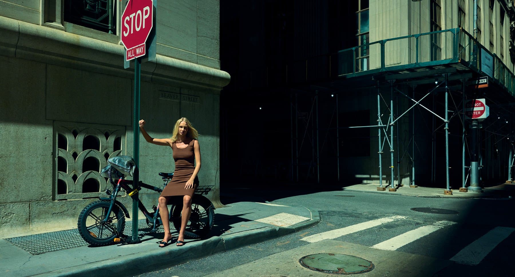 A fashion photograph of a woman in a brown dress with a bicycle on a sunlit New York street corner.