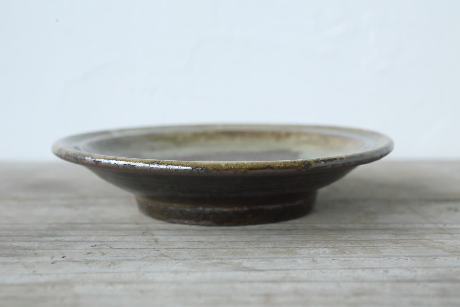 Low side view of a shallow brown-green glazed plate showing its curved rim and raised foot.