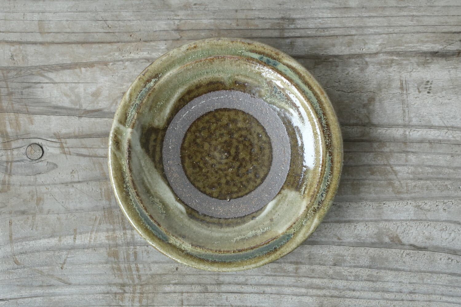 Overhead view of a round plate with green-brown glaze and a grey circular band on weathered wood.