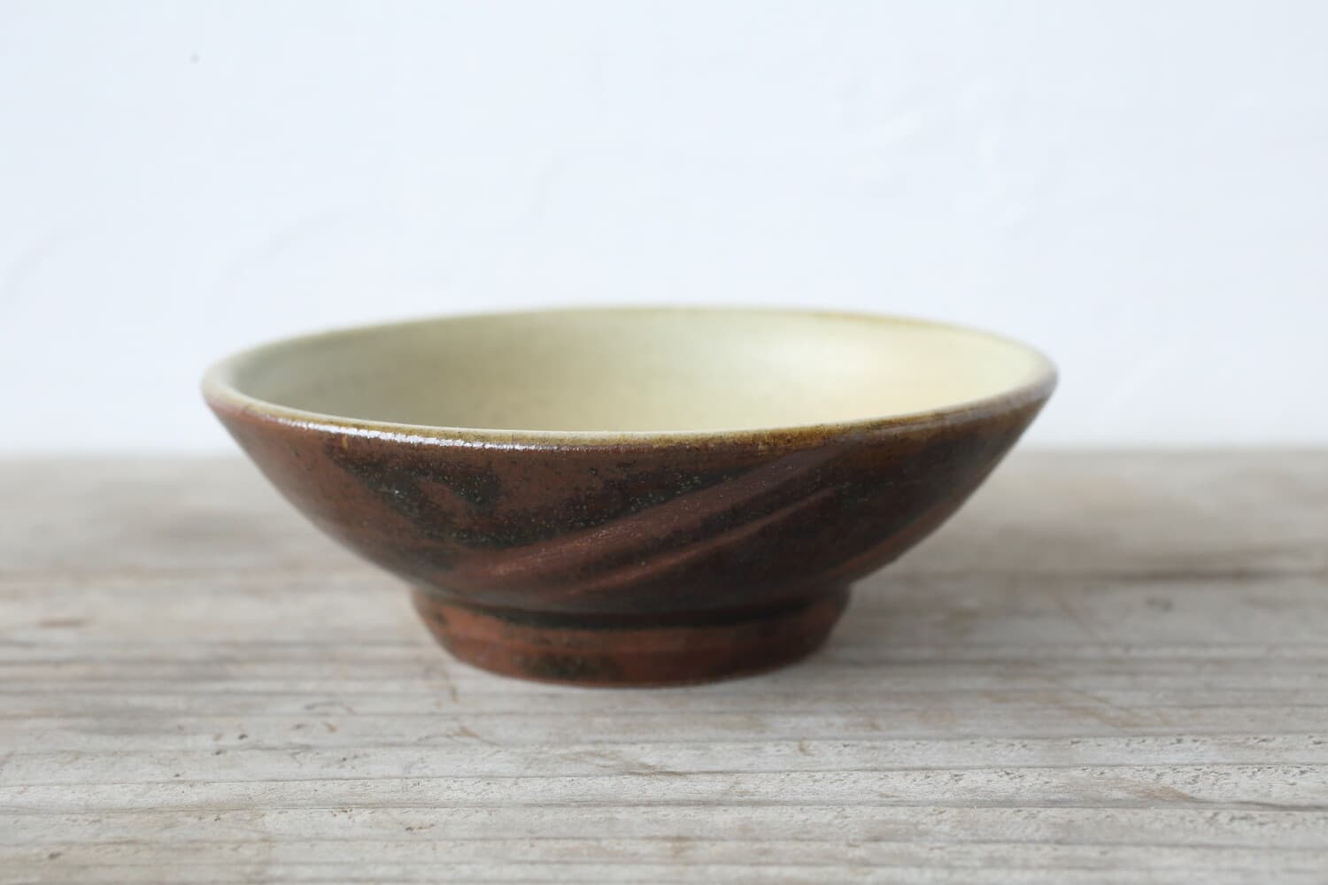 A shallow bowl with a brown exterior, dark curved brush marks and a pale yellow-green interior rests on wood.