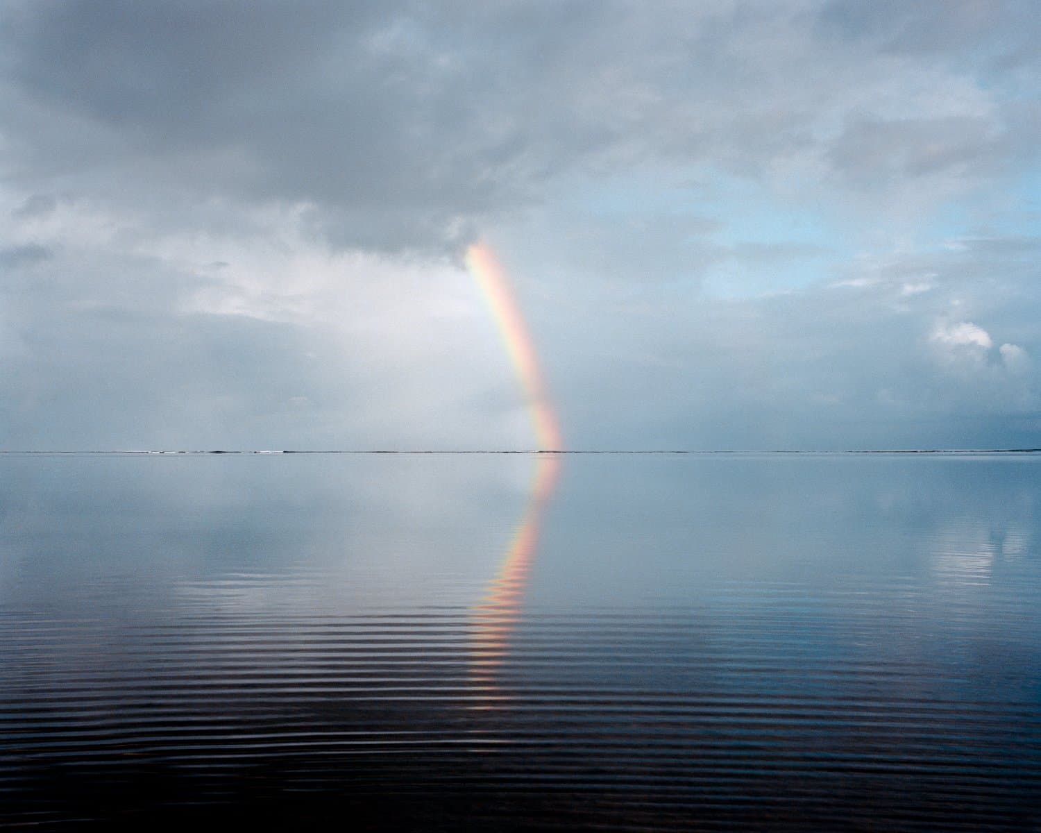 A rainbow reflected in still water under a cloudy sky.