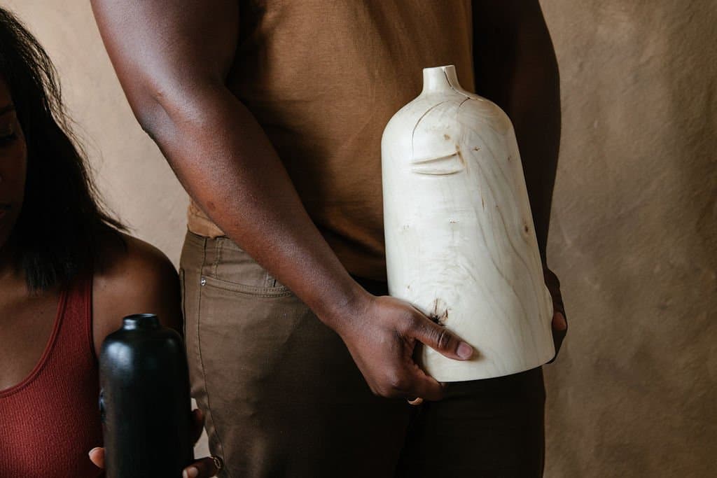 A hand-carved pale wooden Bel vessel from Two Tree Studios, held in a person’s arms beside a smaller dark vessel.