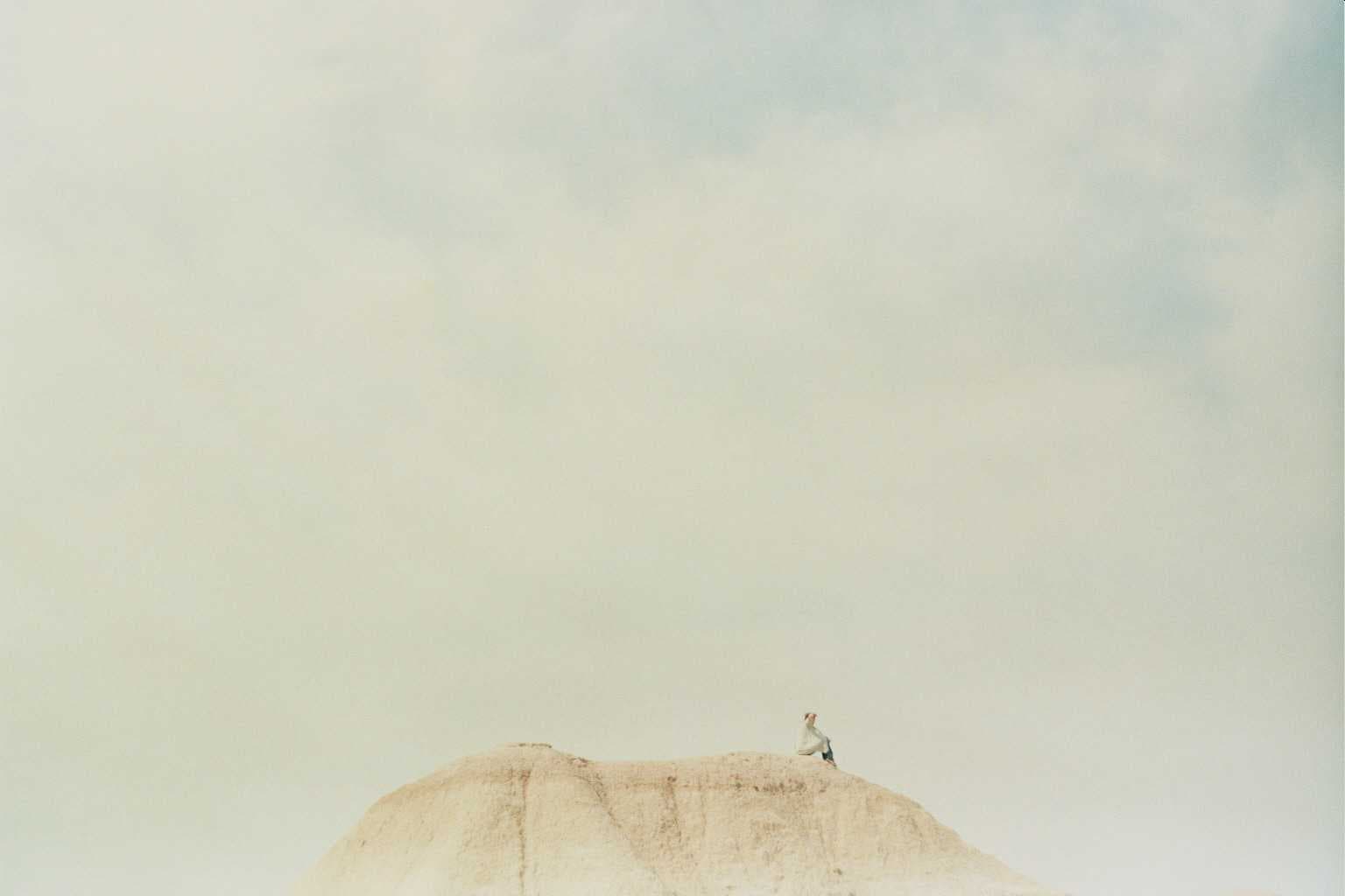 A small distant figure sits on a pale dune beneath a wide, softly toned sky.
