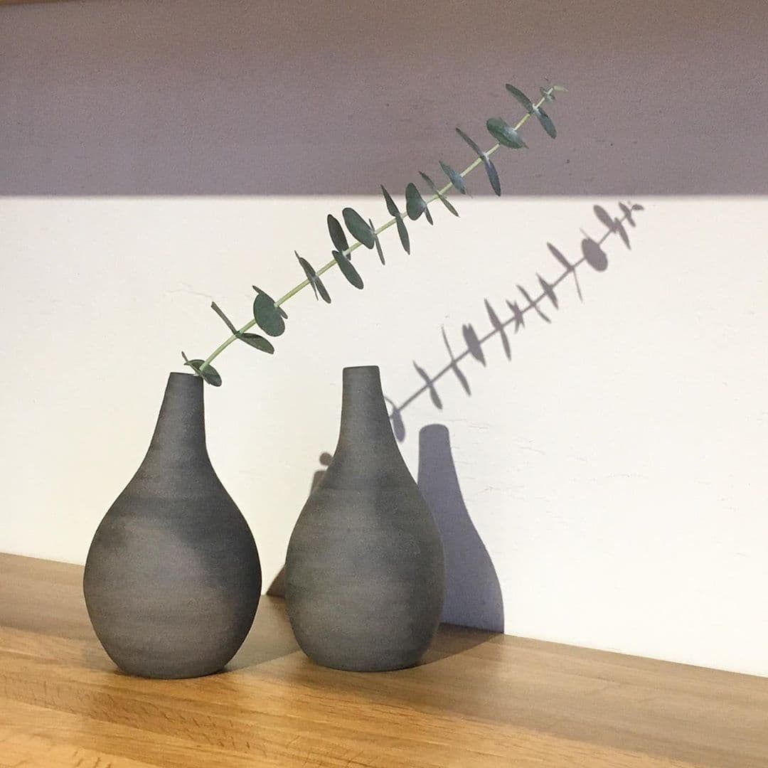Two gray Bulb ceramic vases by Nozomi Fujii on a wooden shelf, one holding a leafy stem.