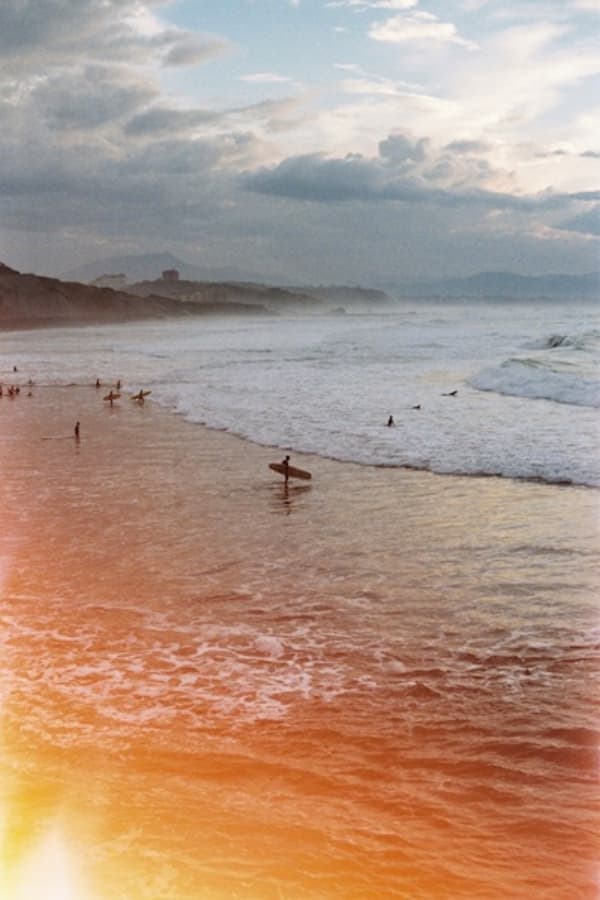 A beach with small distant surfers and a warm orange light flare across the foreground.