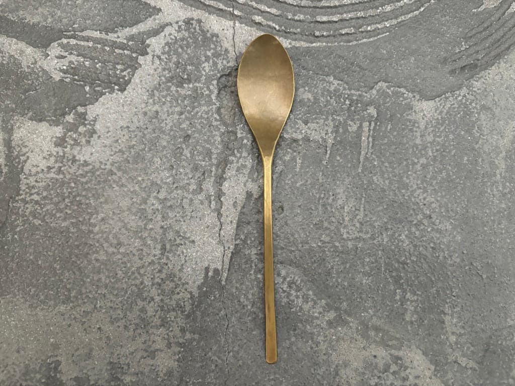 Handmade brass dessert spoon by Tomomi Ishikawa, shown whole against a textured grey surface.