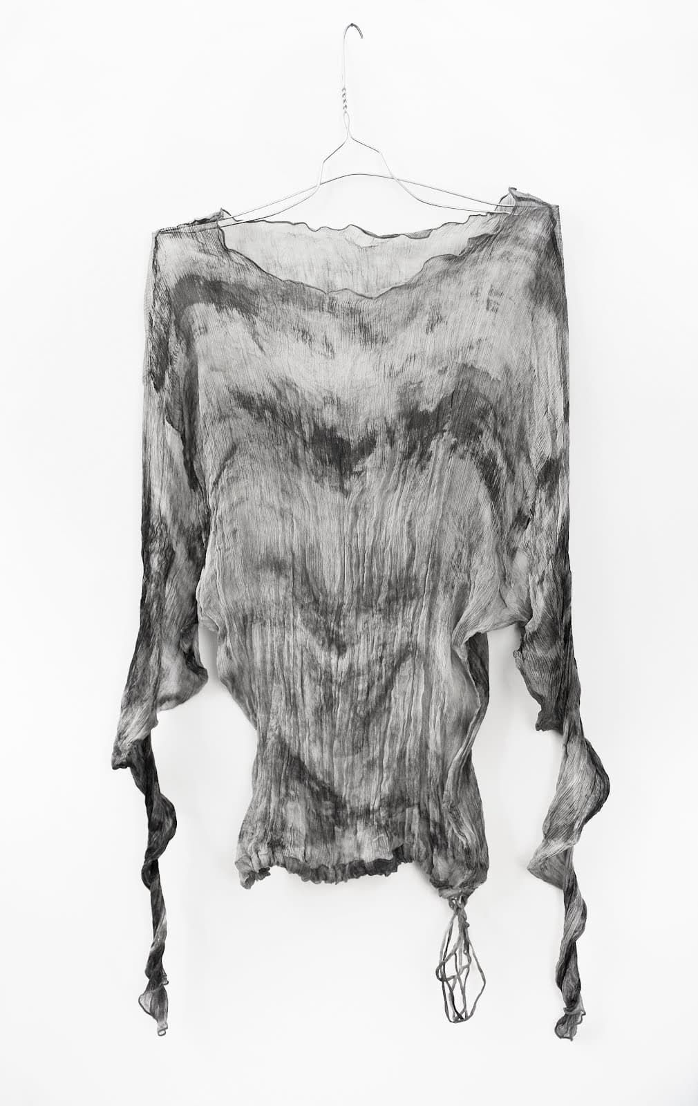 Black-and-white photograph of a textured long-sleeved garment hanging from a wire hanger against a plain pale background.