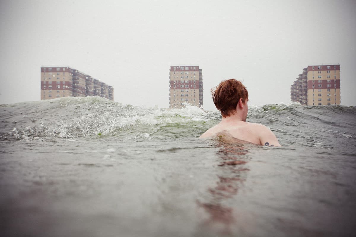 A back-facing swimmer floats in choppy water, with three apartment buildings beyond the waves.