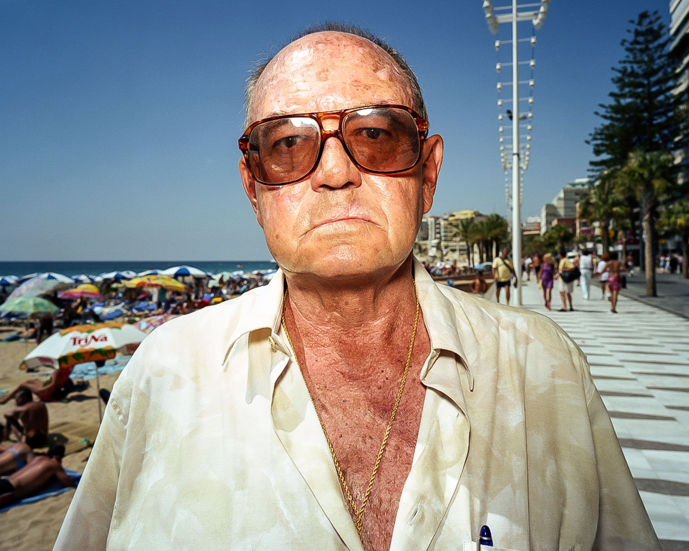 Close portrait of an older adult man wearing sunglasses and a light patterned shirt on a busy seaside promenade.