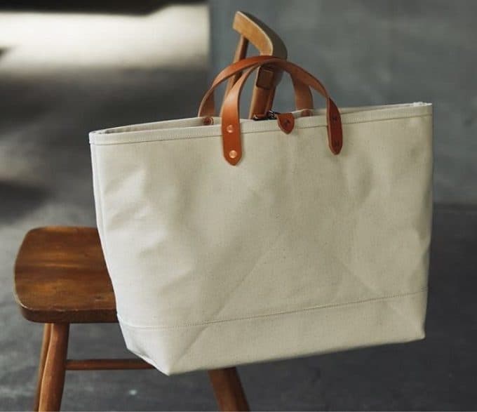 A natural canvas KEESE tote with brown leather handles and metal rivets, resting on a wooden chair.