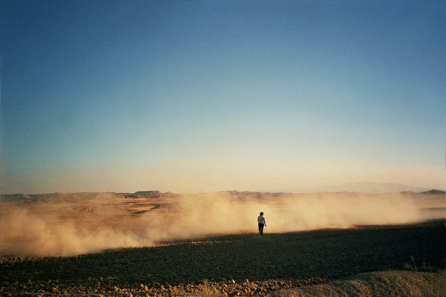 A distant, back-facing figure stands in a dusty desert landscape beneath a clear blue sky.