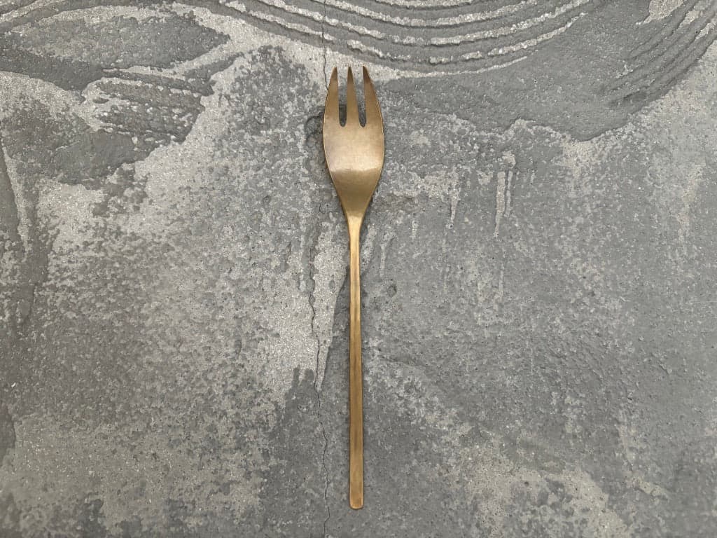 Handmade brass dessert fork by Tomomi Ishikawa, shown whole against a textured grey surface.