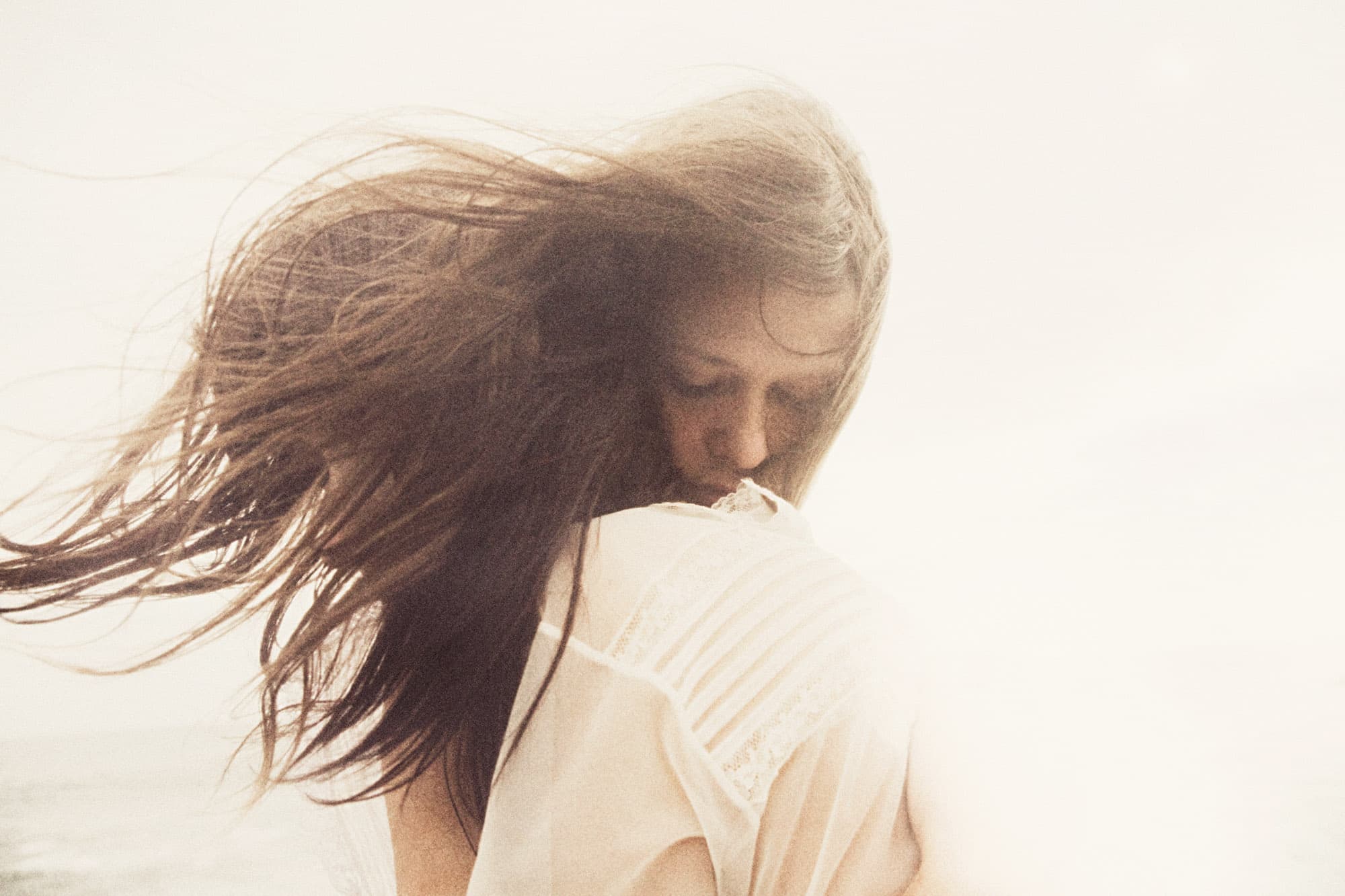 A windblown adult woman in a pale garment looks downward against a bright, softly rendered background.