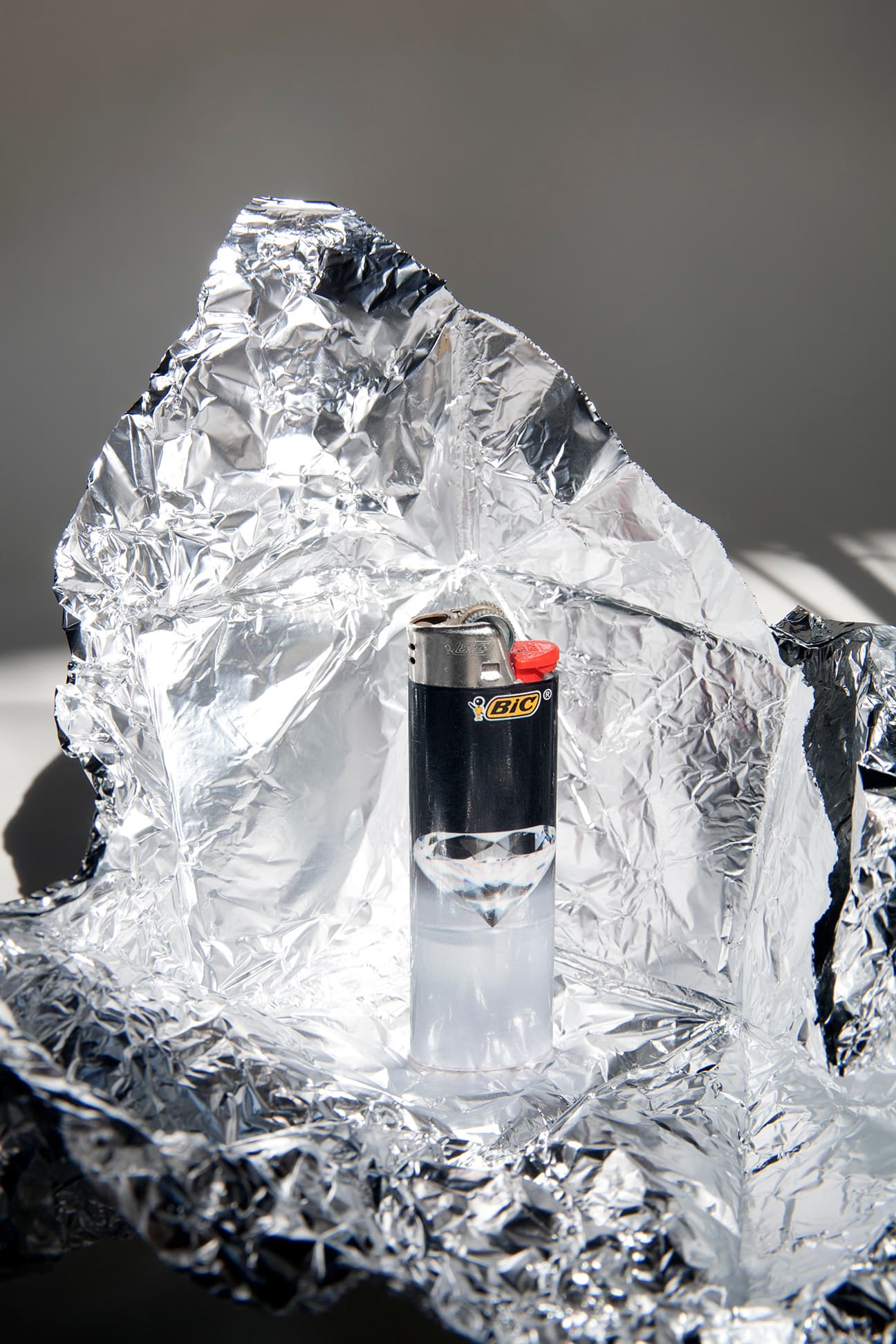 A clear cigarette lighter stands against crumpled reflective aluminium foil.