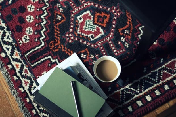 Notebooks, a pen, newspaper and coffee cup rest over a patterned rug beside a chair.