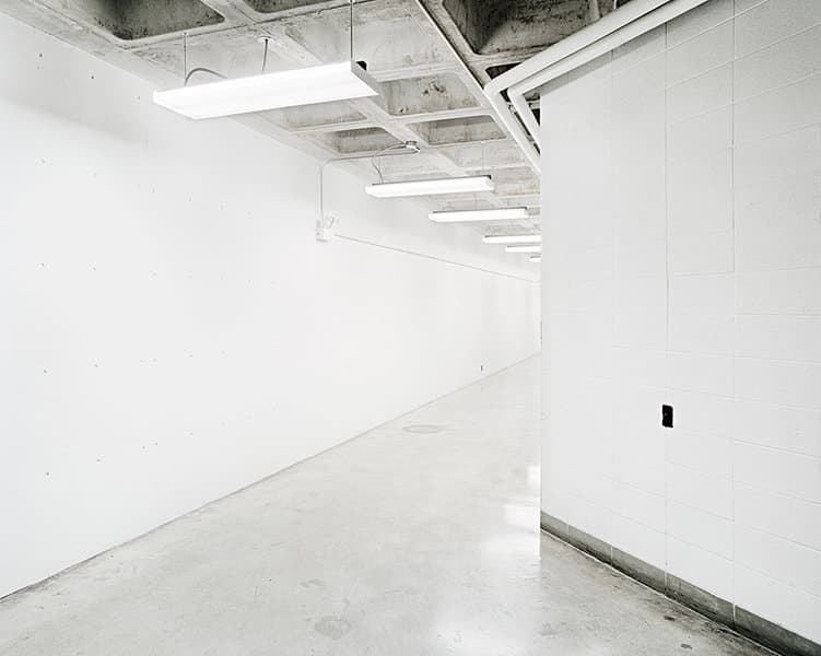 A bright empty corridor curves around a tiled white wall under exposed ceiling beams.