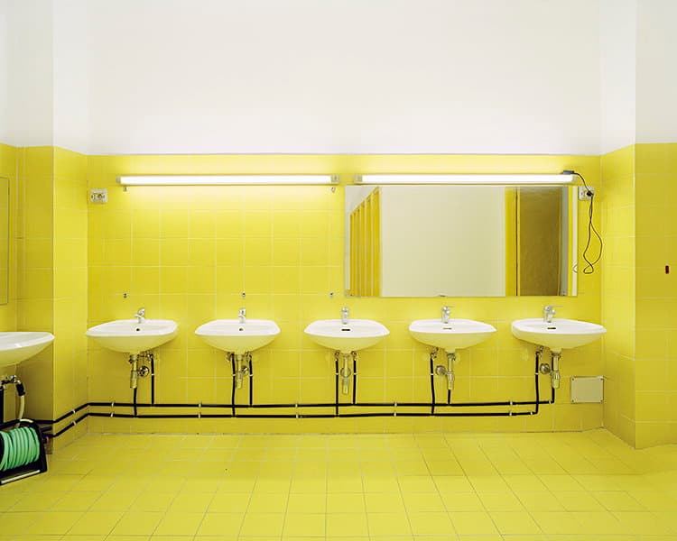 White basins line a bright yellow tiled washroom beneath fluorescent lights and a mirror.