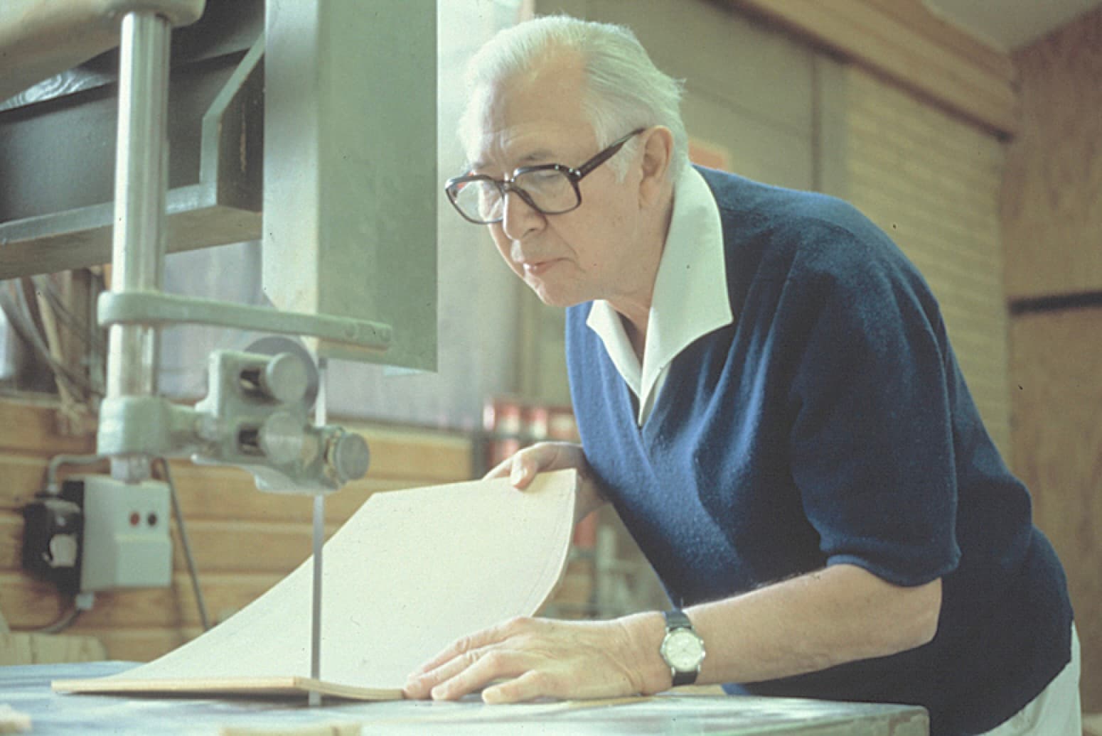 Hans J. Wegner guides a shaped piece of wood through a bandsaw in a workshop.