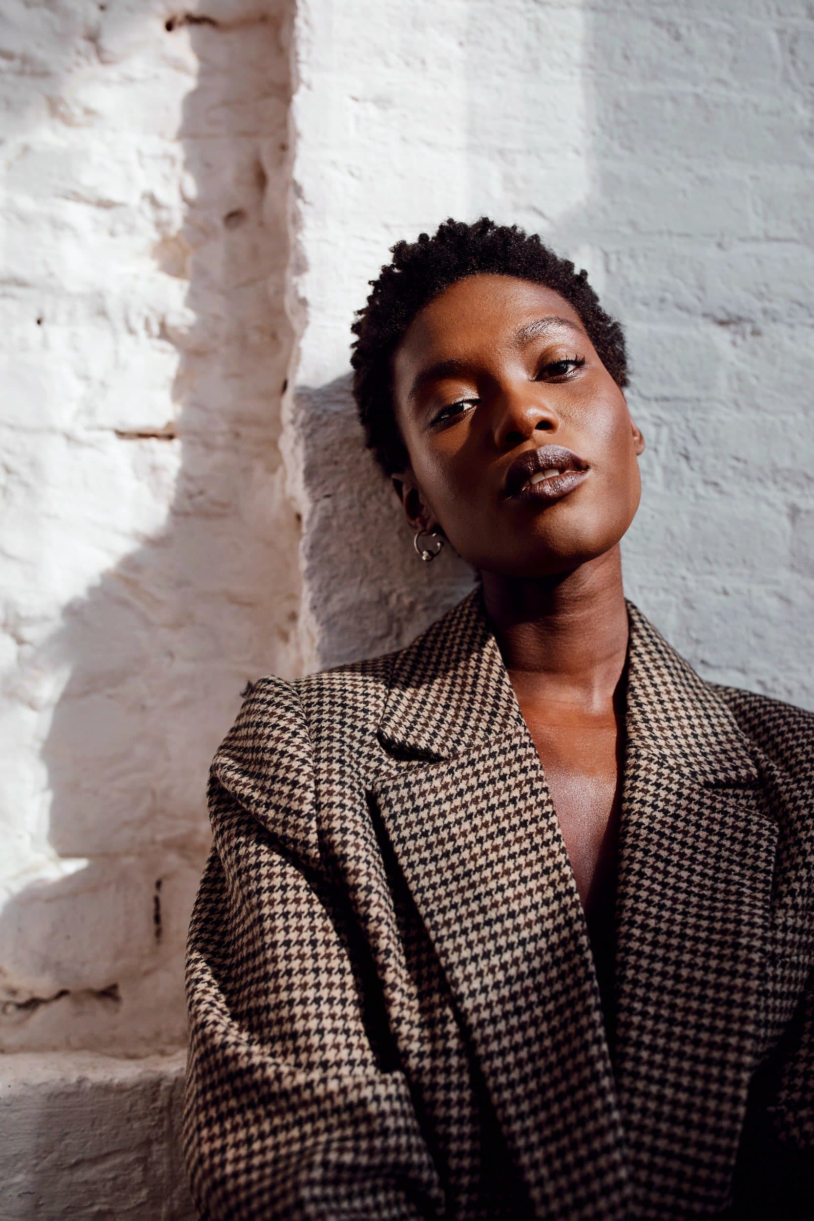An adult woman in a houndstooth jacket leans against a white brick wall in directional sunlight.