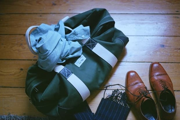A dark green holdall, folded clothes and brown leather brogues on a wooden floor.