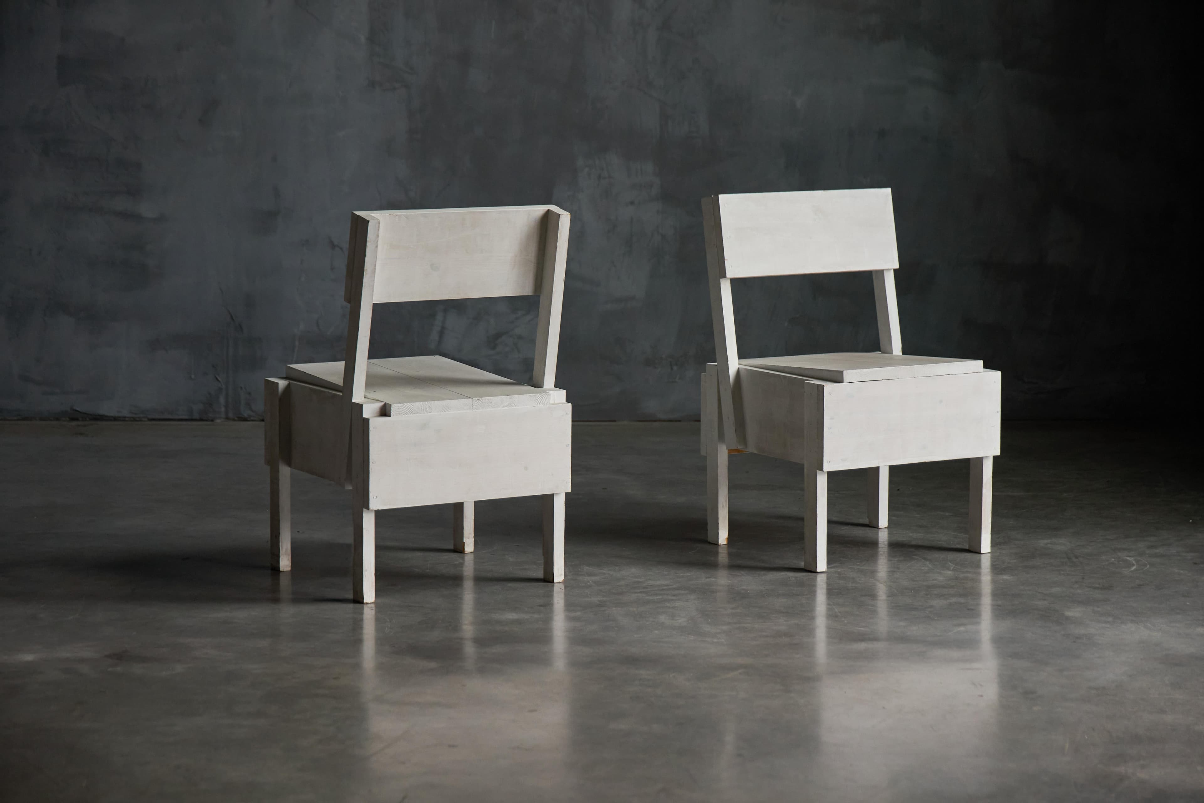 Two pale wooden Sedia 1 chairs stand on a concrete floor against a dark grey wall.