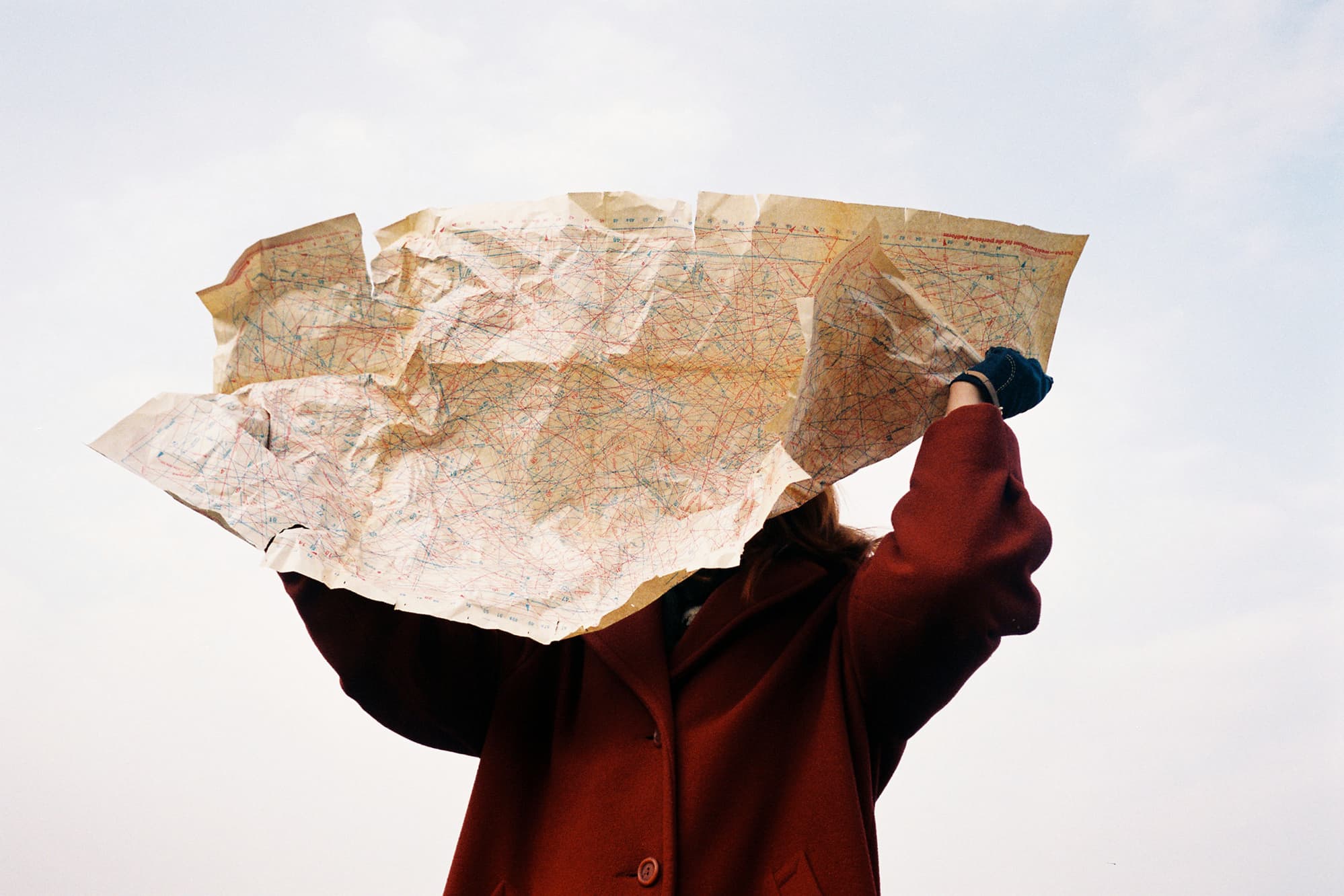 A burgundy-coated figure holds a large crumpled sewing pattern in front of their face against a pale sky.