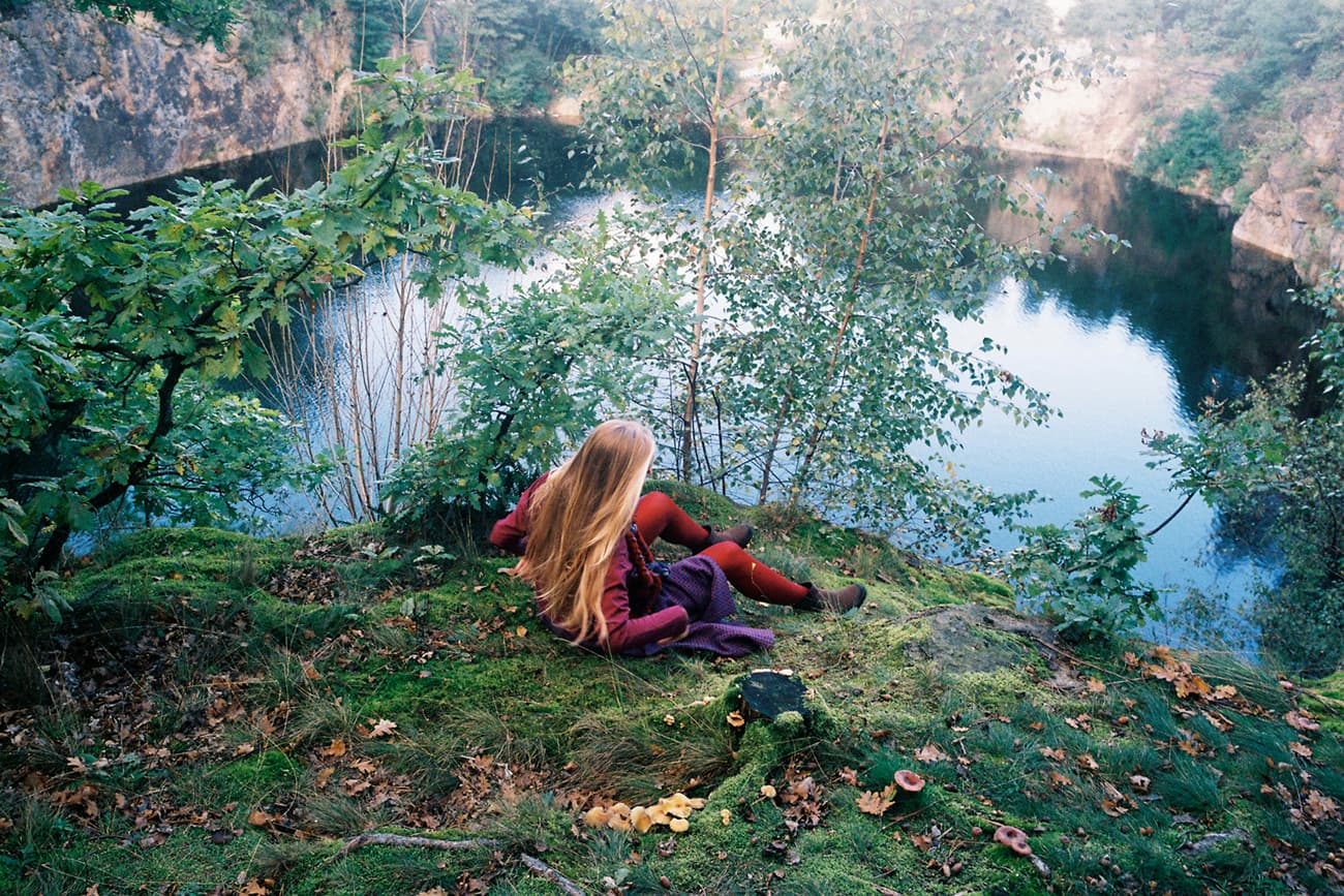 A long-haired, red-clad figure sits on a mossy ledge above a still wooded lake.