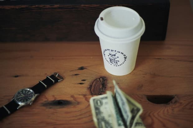 A paper coffee cup, wristwatch and folded cash lie on a wooden tabletop.