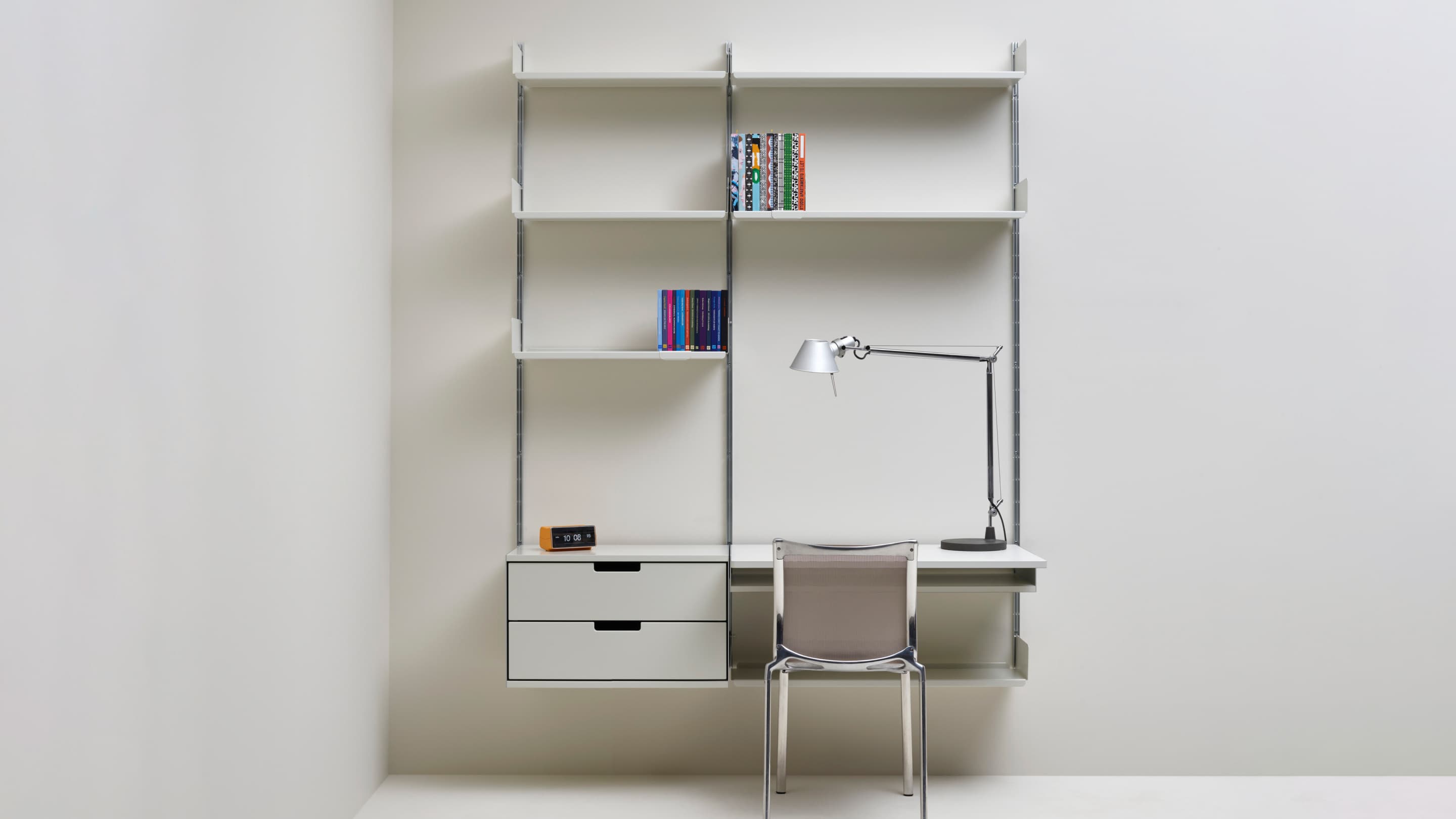 An off-white wall-mounted shelving system with two drawers, a desk, books and a task lamp, shown with a chair.