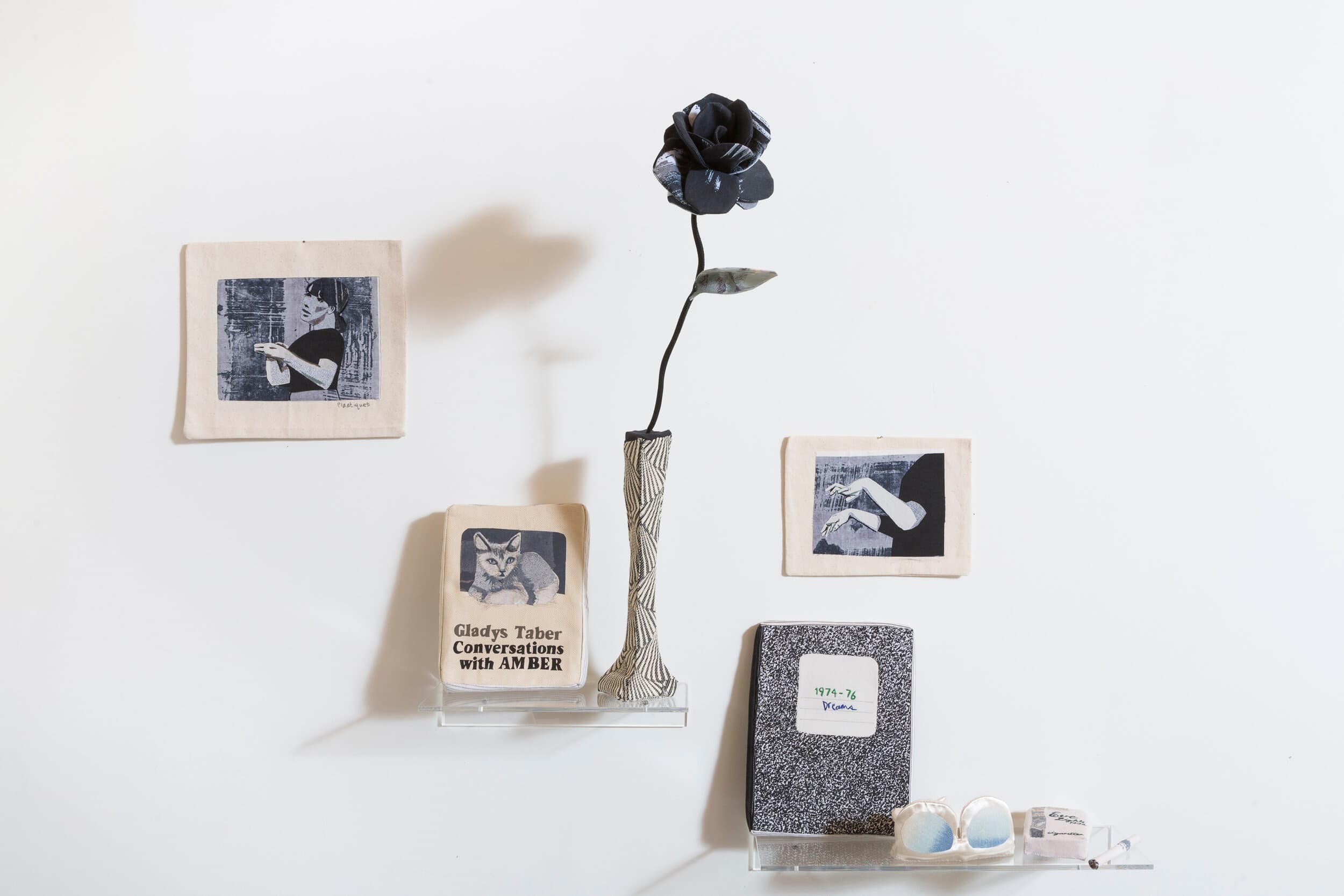 Fabric objects arranged on clear wall shelves, including a book, notebook, flower in a vase, glasses and illustrated cloth panels.