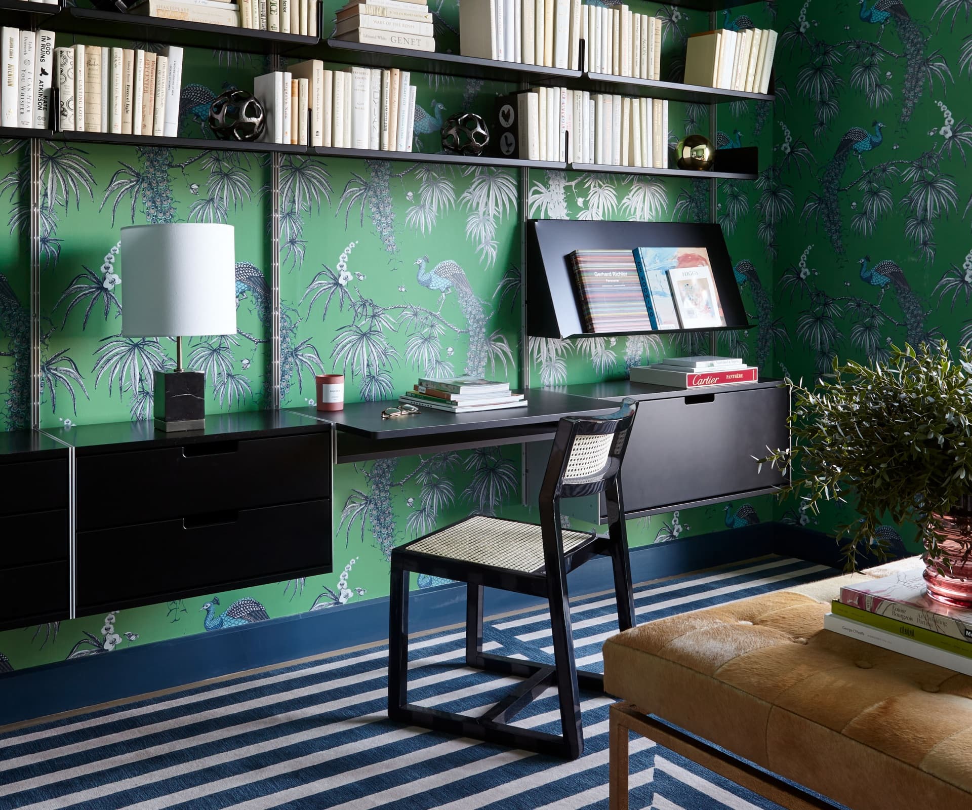 Black 606 shelving, drawer cabinets and a desk against green patterned wallpaper, with books, a lamp and a chair.