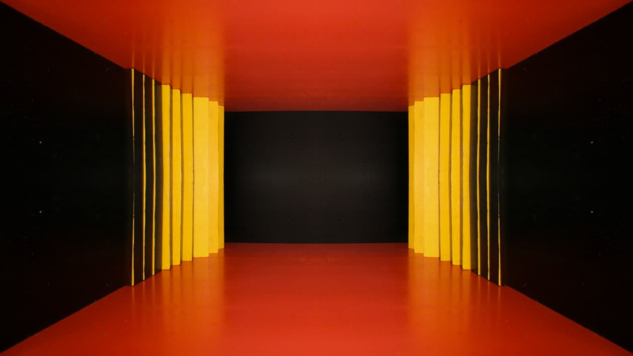 Yellow and black wooden strips line the sides of an orange-red geometric tunnel with a dark back wall.
