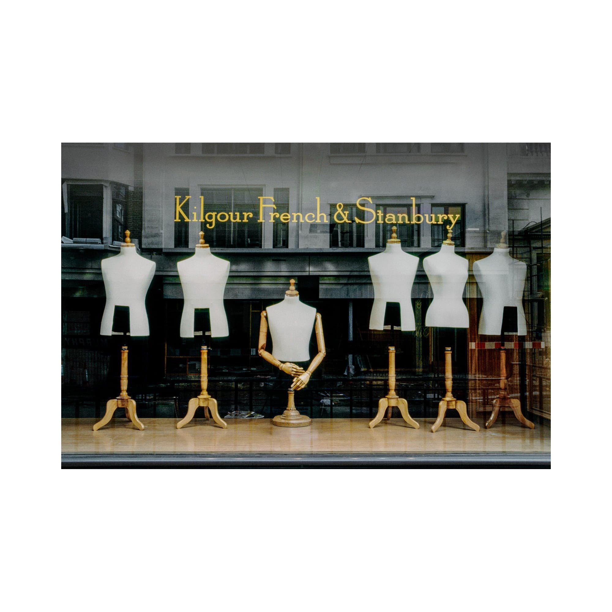 Six empty dress forms in the Kilgour French & Stanbury shop window on Savile Row, beneath gold lettering and reflections of London buildings.