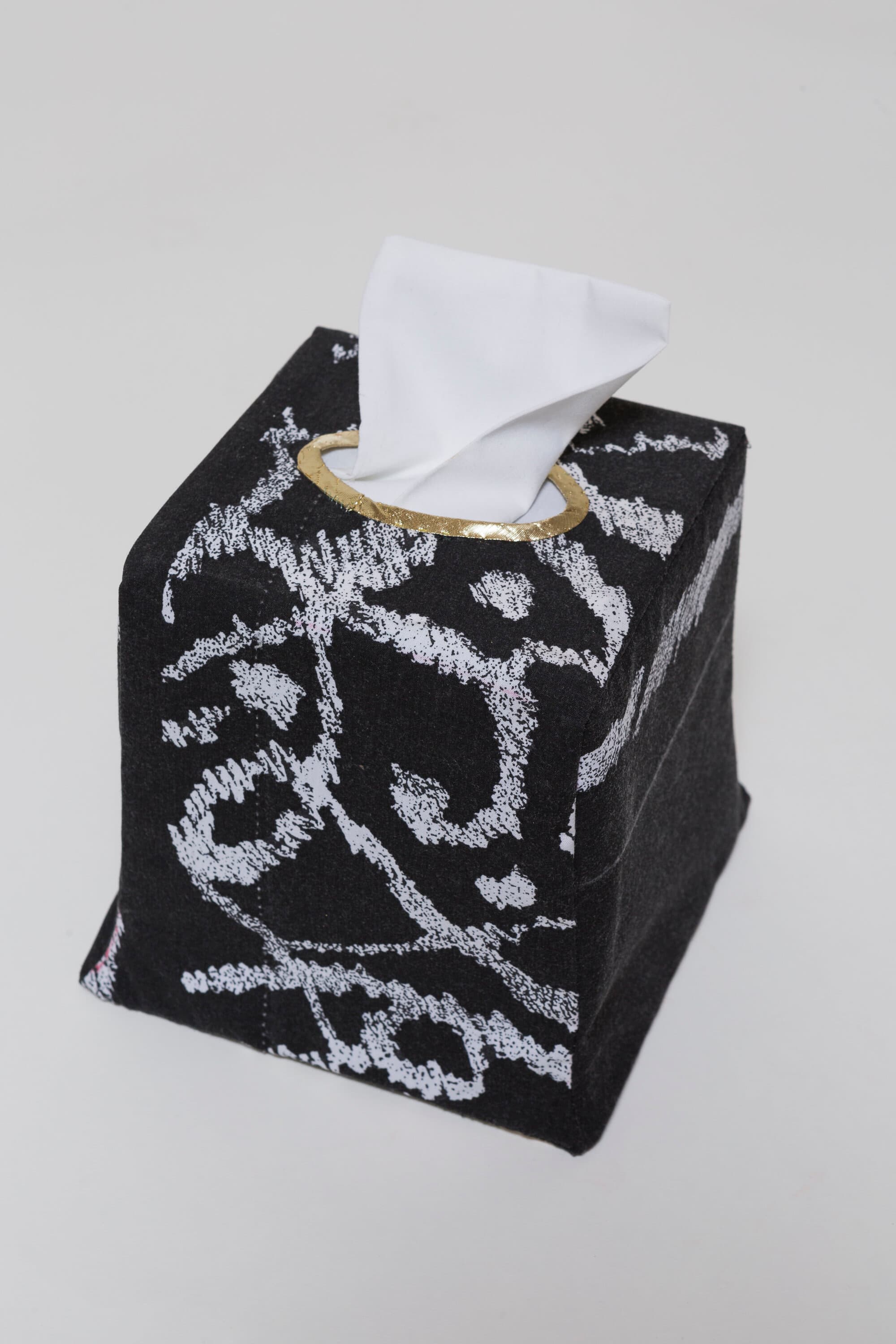 A black-and-white patterned fabric tissue box with a gold-edged opening and a white cloth tissue protruding from the top.