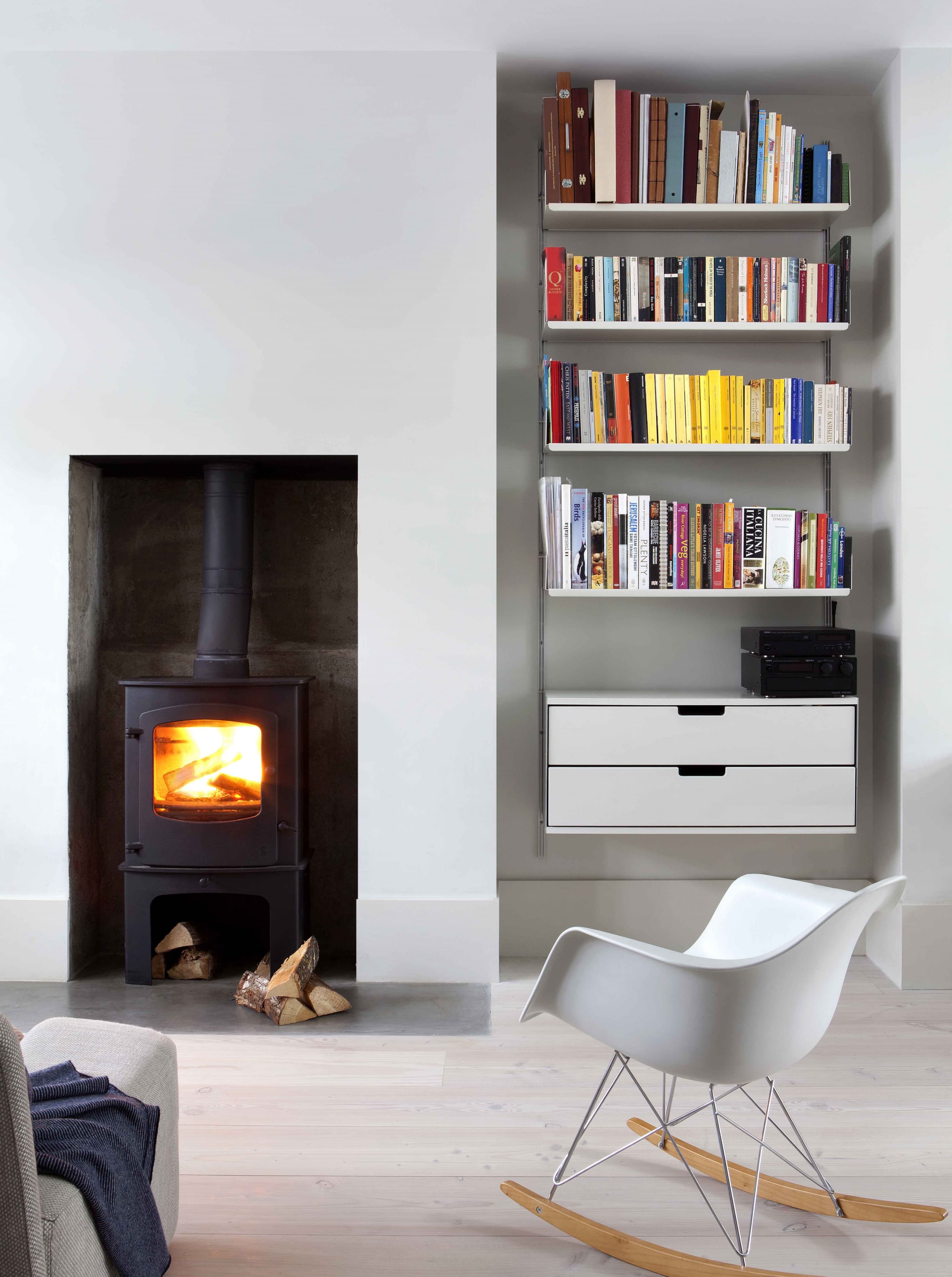 Off-white 606 shelves filled with books above two drawers in an alcove beside a wood-burning stove.